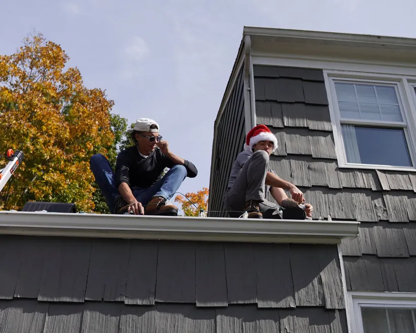 Danny and Joey on a roof clipping holiday lights to the gutter, one wearing a Santa hat