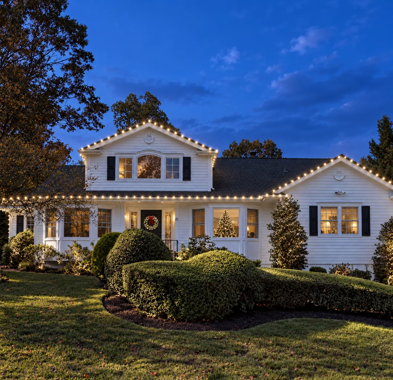 A white colonial house at dusk outlined in warm white holiday lights, with a lit wreath on the door